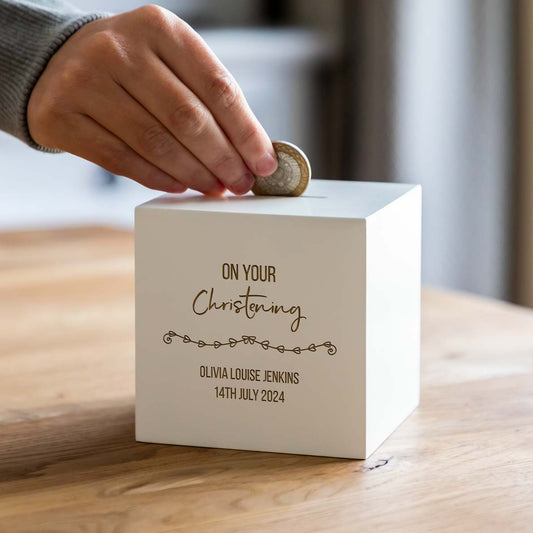 A personalised white wooden money box with 'On Your Christening' text engraved on it, with a name and date below in the image.