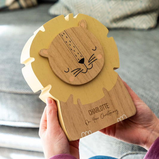 A personalised wooden lion-shaped money box engraved with a child's name and 'On Your Christening' text, being held by a person.