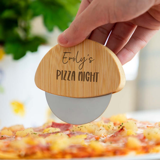A person holding a wooden pizza cutter engraved with a name and 'Pizza Night' text, over a pizza.