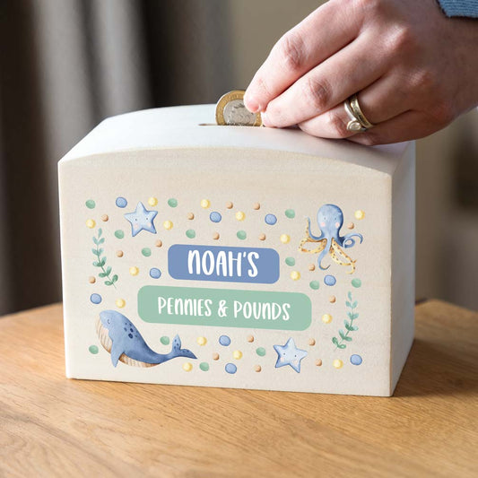 Children's money box personalised with any text, printed with sea-themed illustrations on a wooden surface.