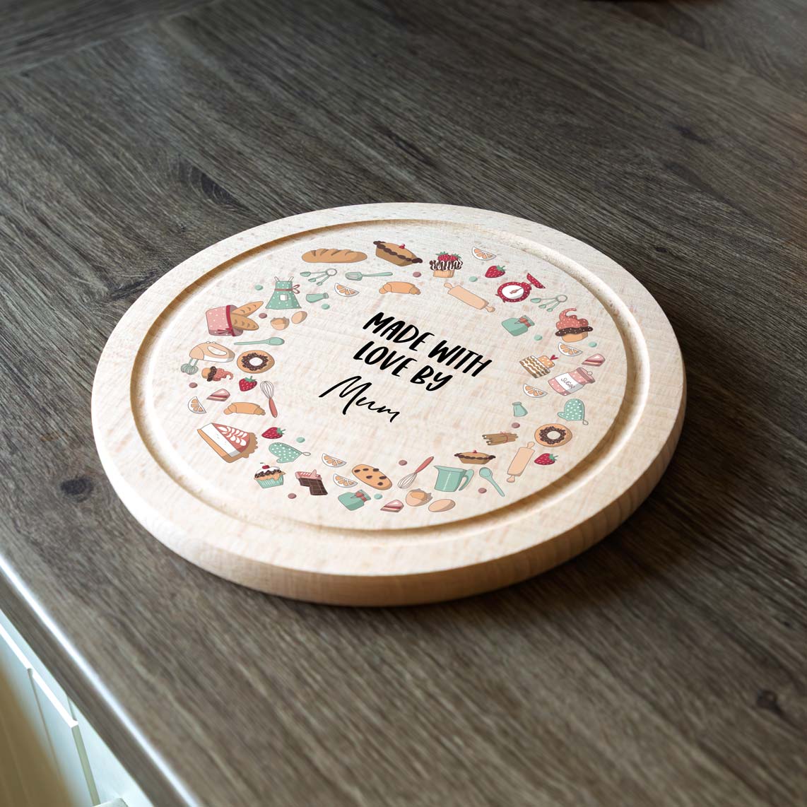Round wooden cake board with colourful baking illustrations and 'Made with Love by' text on a wooden surface.