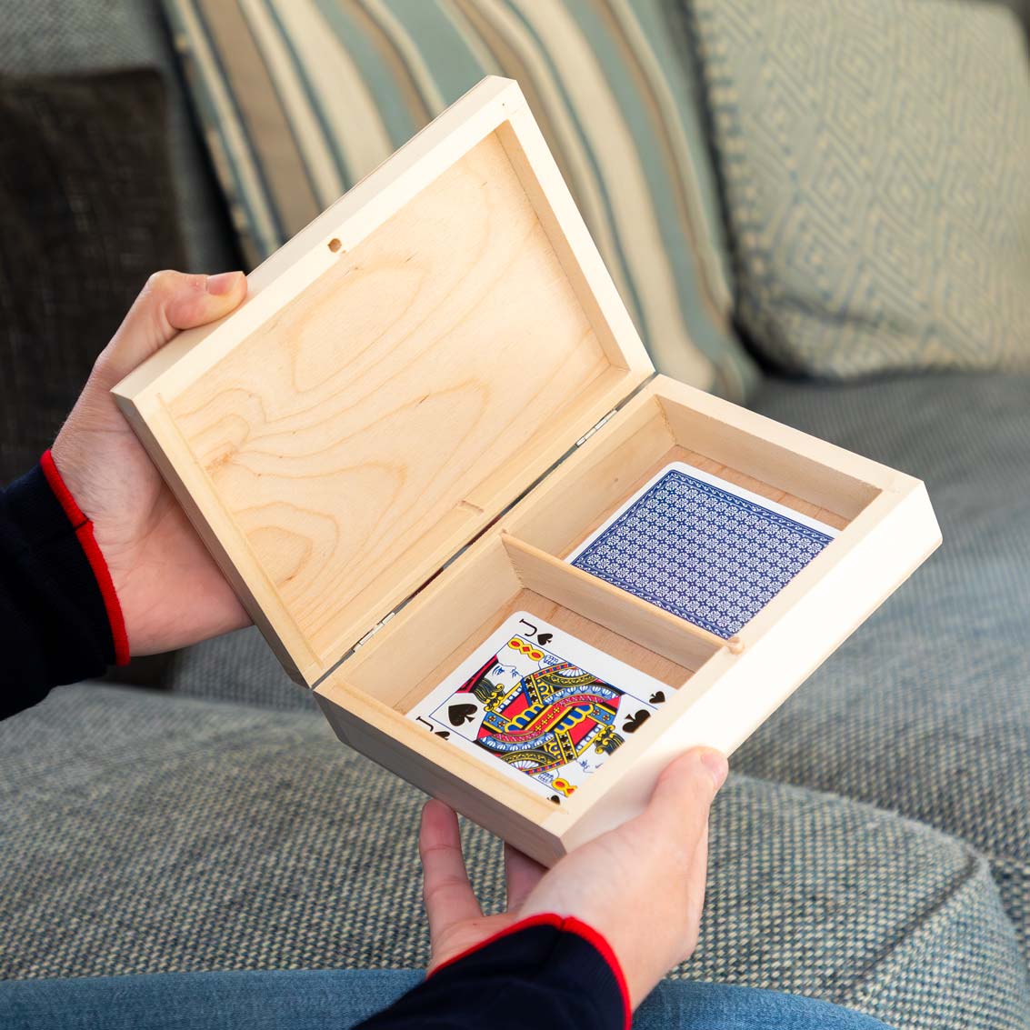 Wooden playing cards box with playing cards inside, held by a person on a couch.