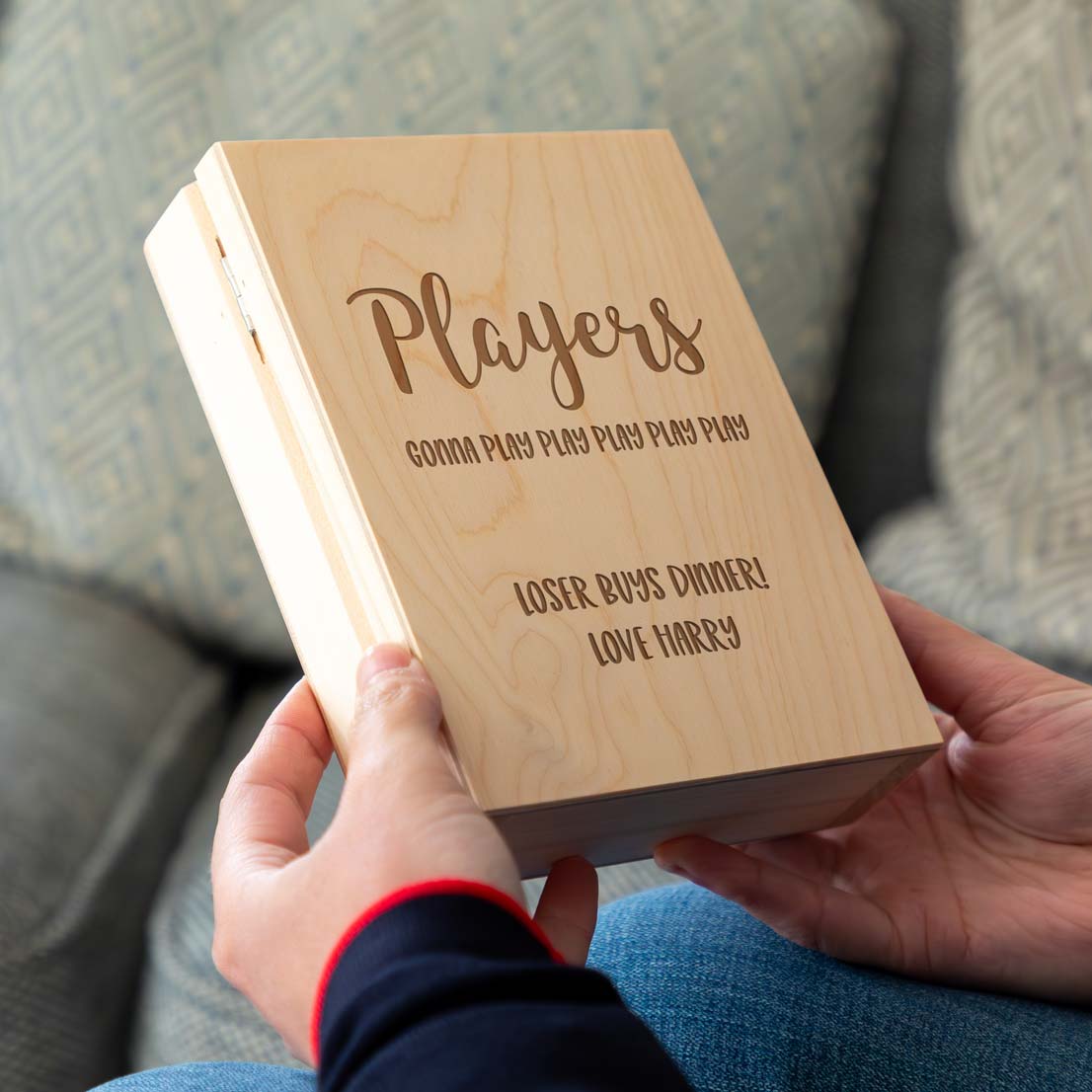 Wooden playing cards holder box with engraved text held by a person, sitting on a textured surface.