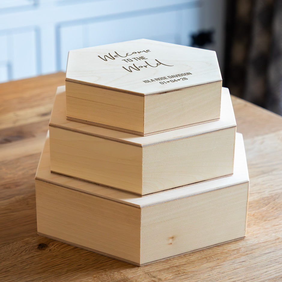 Stack of wooden hexagon keepsake boxes with 'Welcome to the World' text on a wooden surface.