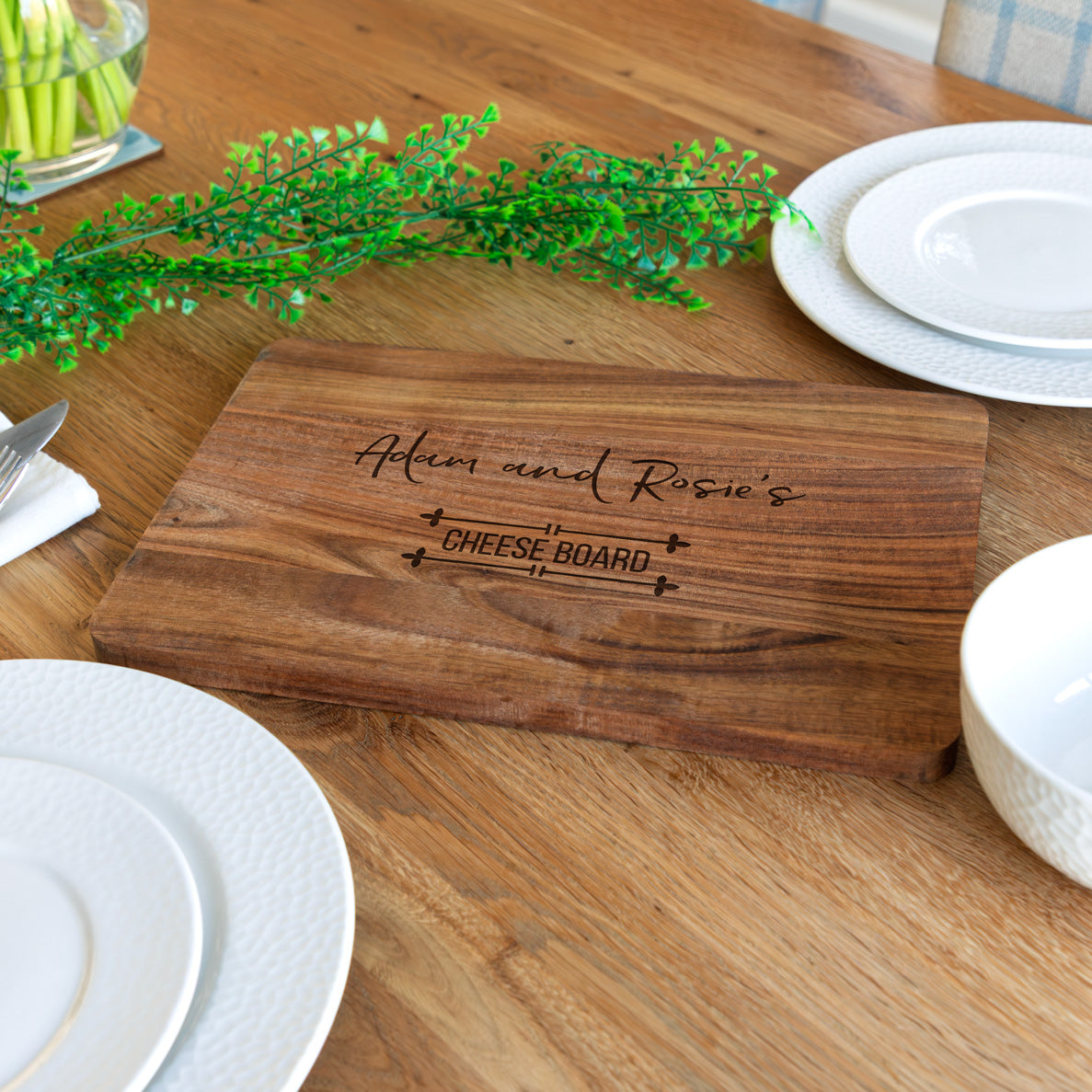 Wooden personalised cheese board with engraved text on a wooden table with plates and greenery.