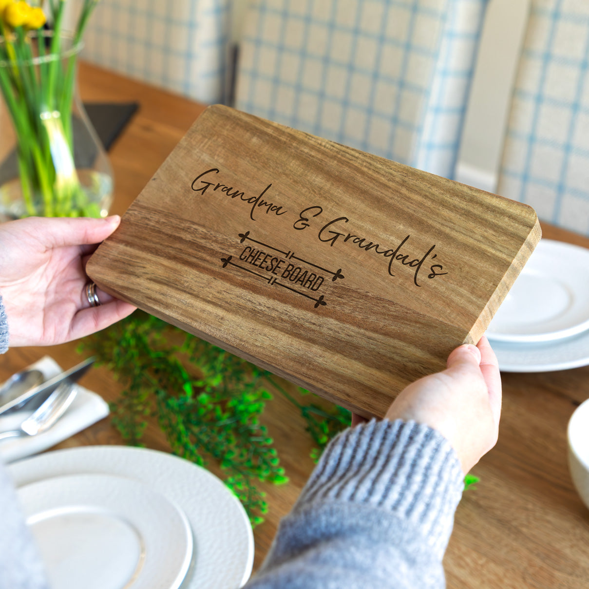 Wooden cheese board personalised with 'Grandma & Grandad's' held by a person on a dining table.