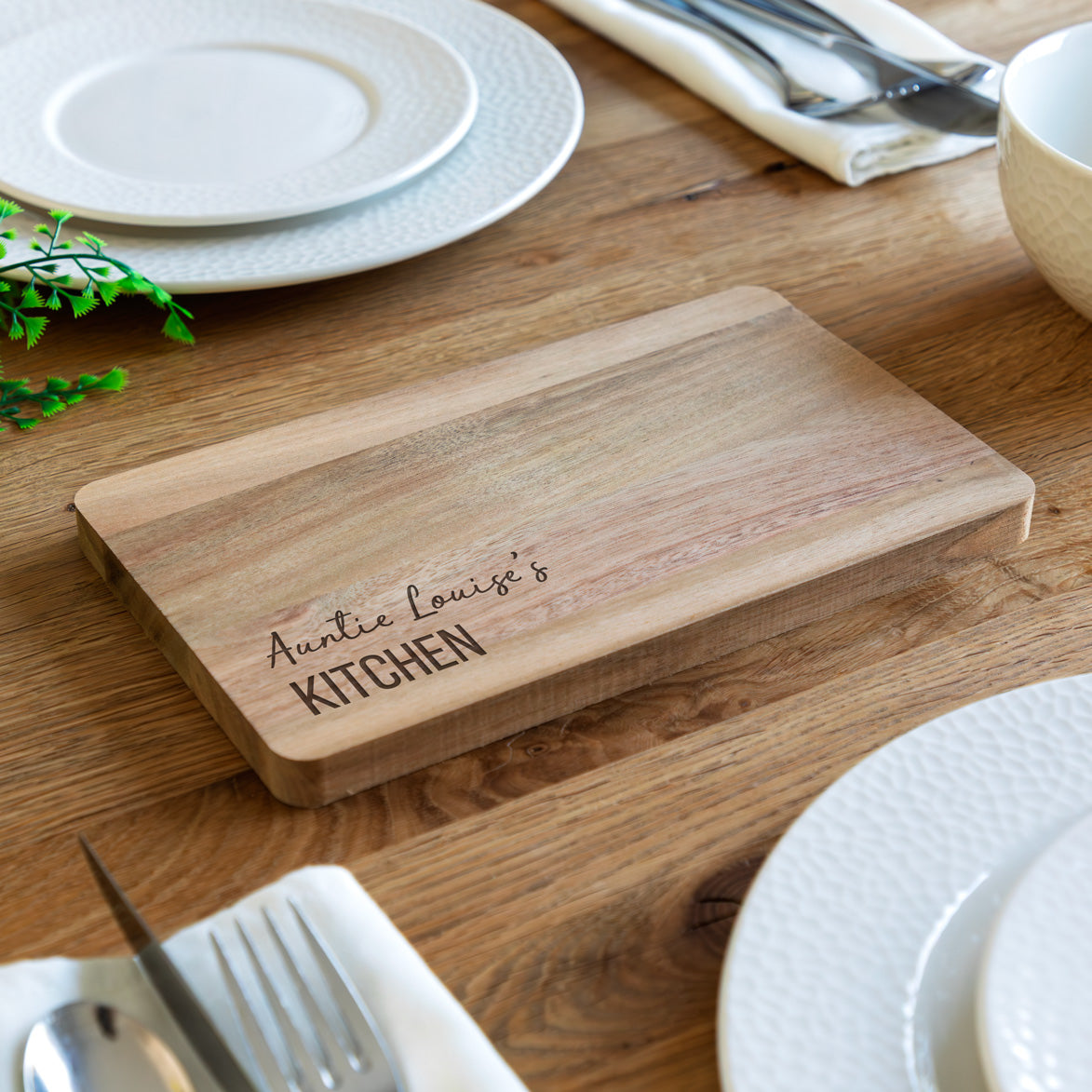 Wooden chopping or serving board with engraved personalised text on a wooden table with plates and utensils.