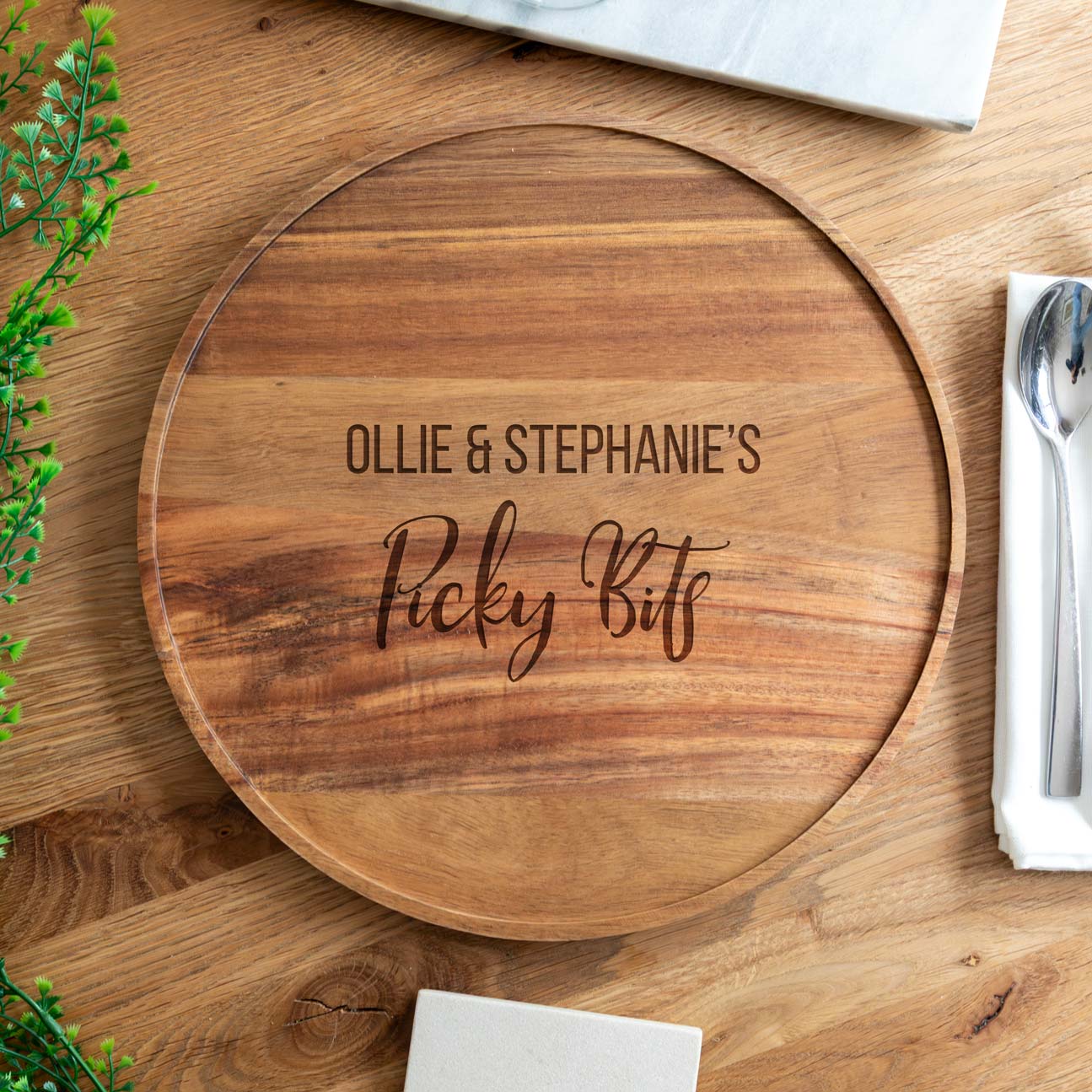 Acacia wood round serving board engraved with the personalised names and 'picky bits' text, on a wooden table.