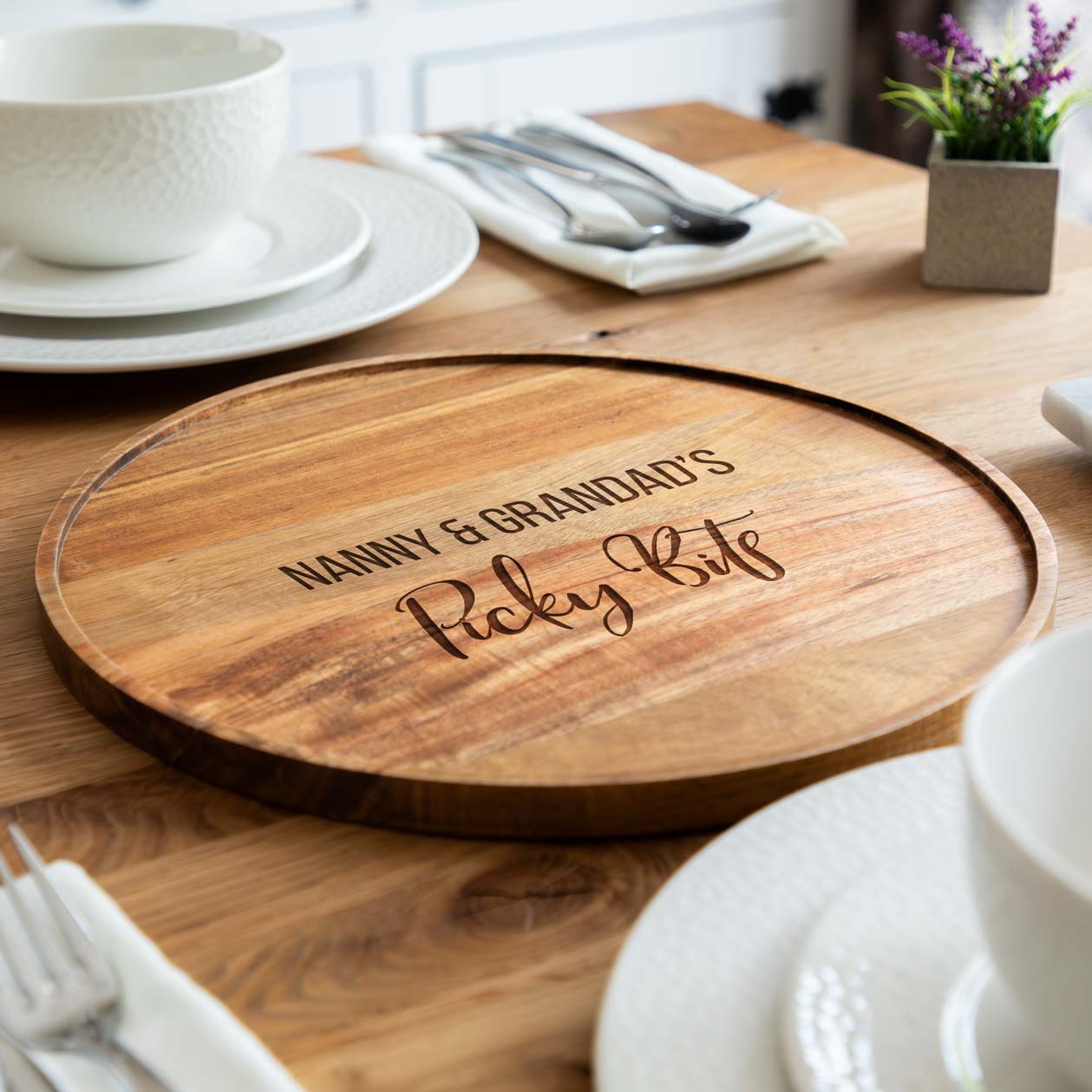 Acacia wood round serving board engraved 'Picky Bits' text, on a dining table setting.