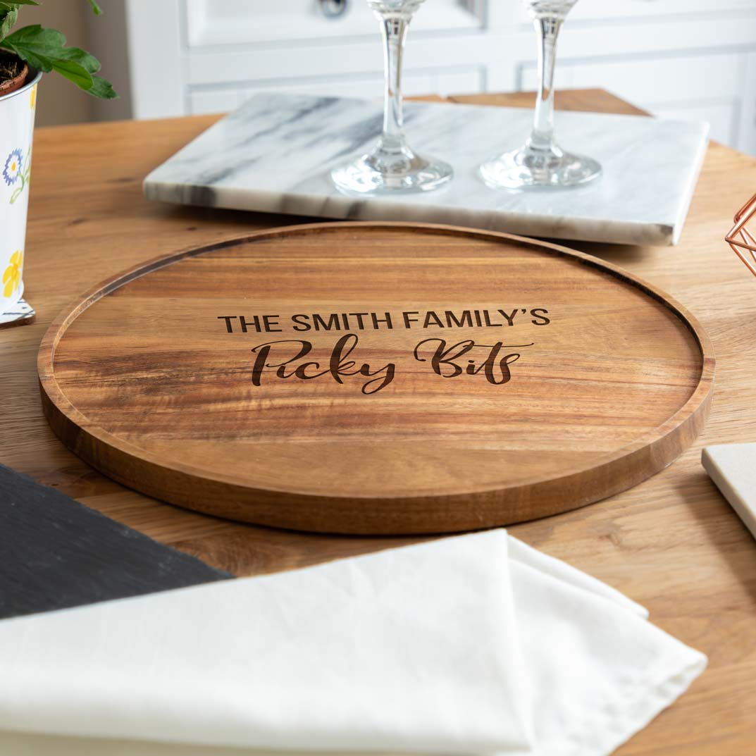 A round wooden serving board engraved with the personalised family name and 'Picky Bits' text, on a dining table.