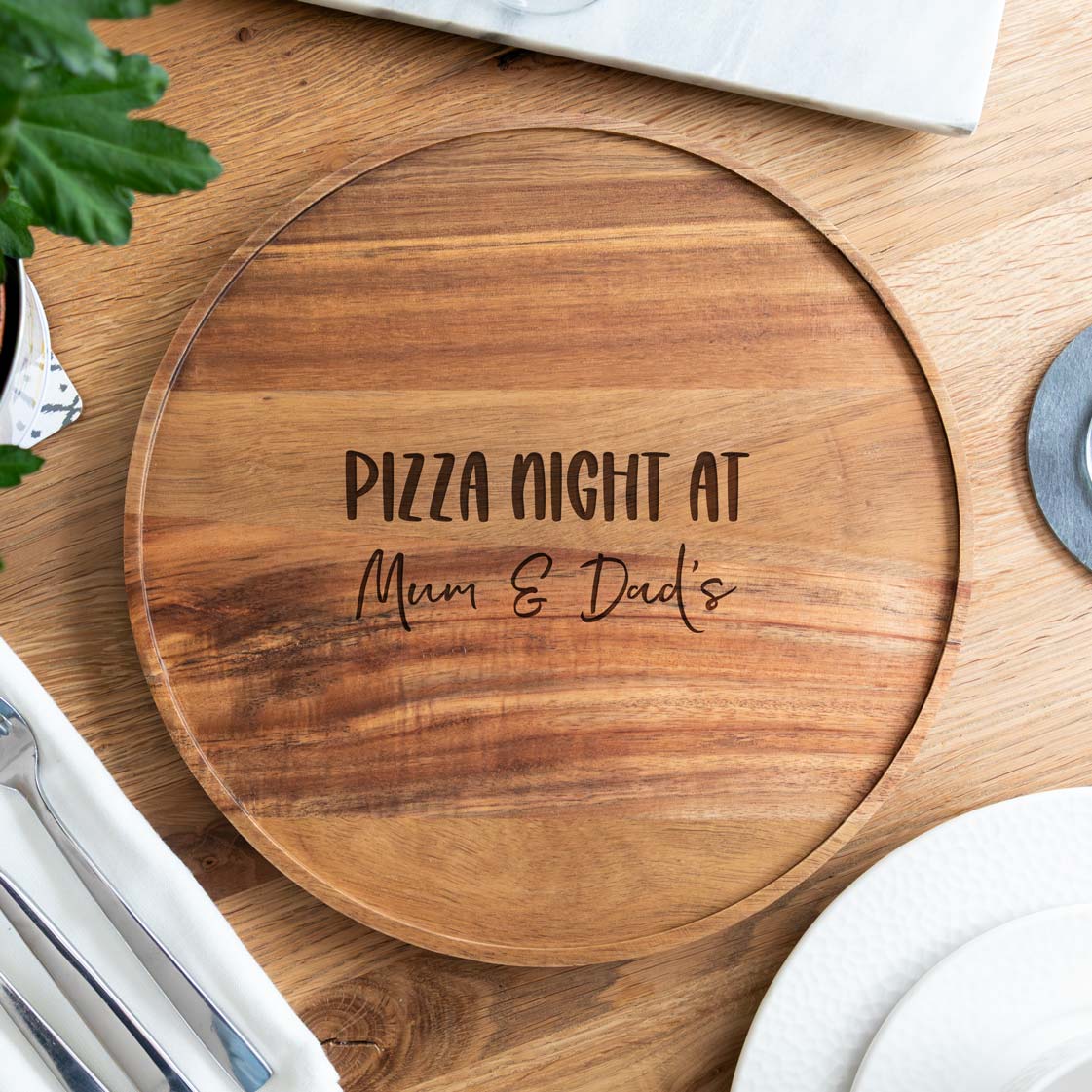 Wooden pizza serving board engraved with 'Pizza Night at' text followed by the personalised names, on a wooden table.