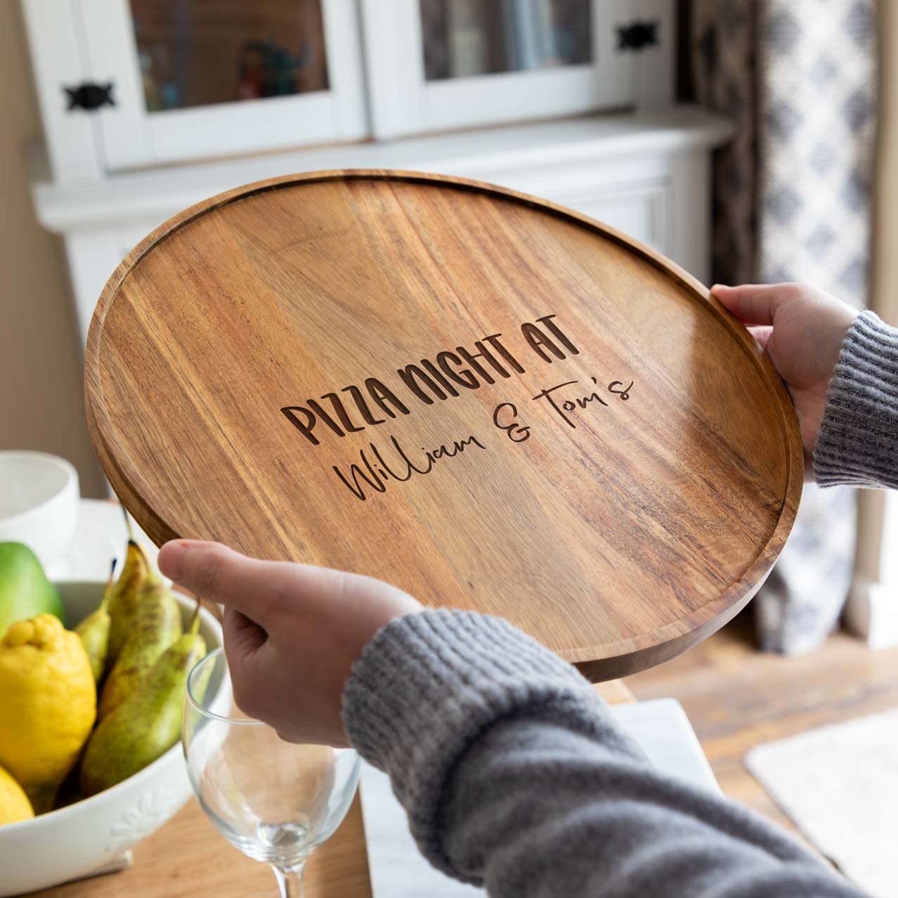 A person holding a wooden pizza board laser-engraved with 'pizza night at' text with the personalised names.
