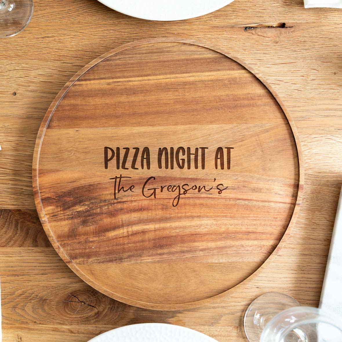 Wooden pizza serving board engraved with 'Pizza Night at' followed by a family name, on a wooden table.