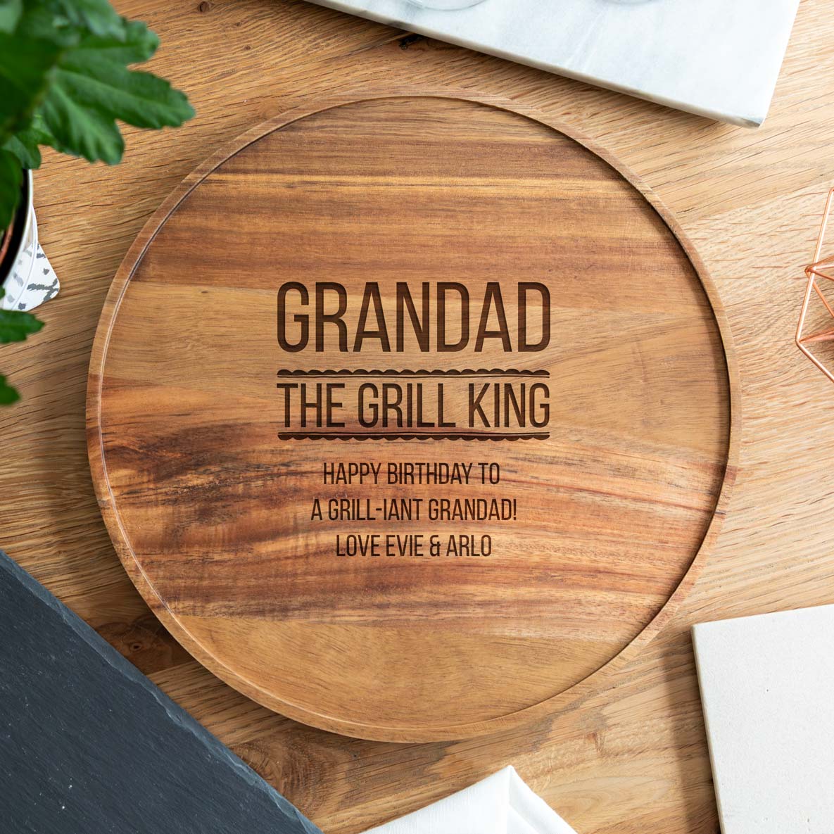 Acacia wood serving board with engraved 'The Grill King' text and a personalised name and message, on a wooden surface.