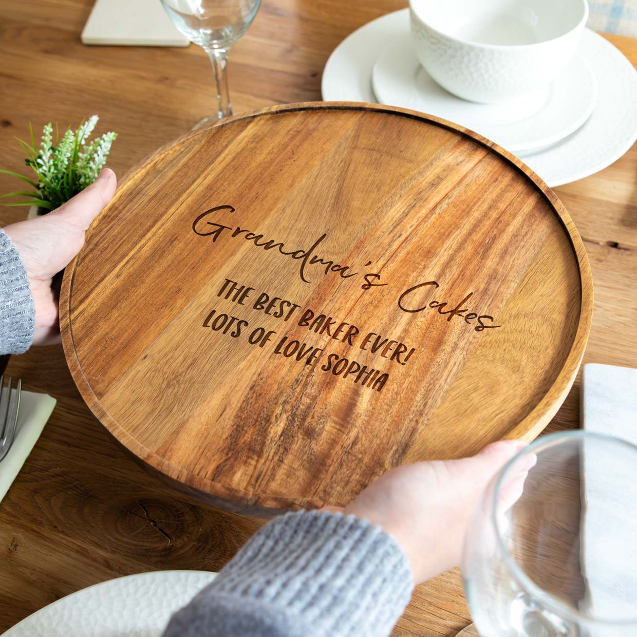 A person holding a wooden acacia cake board with engraved text on a table setting.