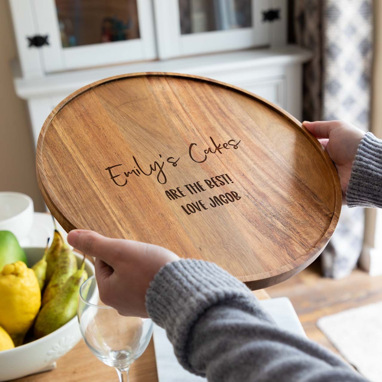A person holding a wooden cake board with engraved personalised text.
