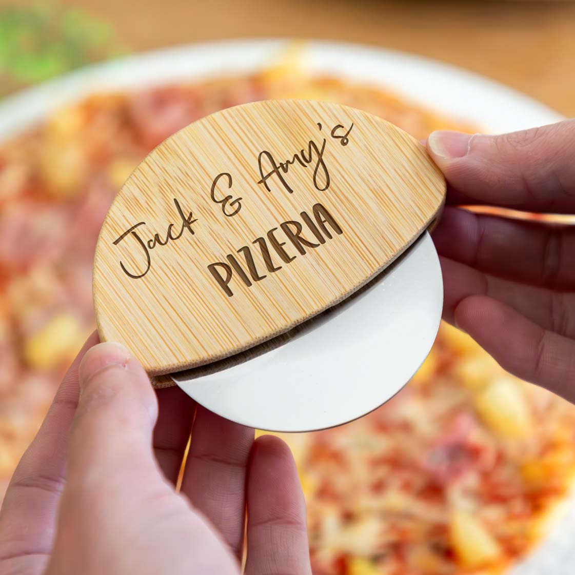 A person holding a wooden pizza cutter engraved with two names and 'Pizzeria' text, over a pizza.