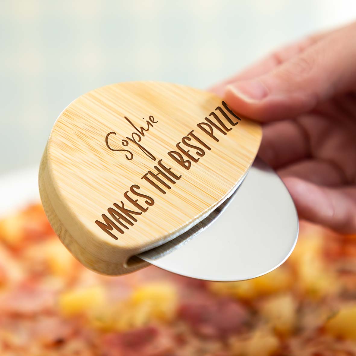 A person holding a wooden pizza cutter engraved with a personalised name and 'makes the best pizza' text.