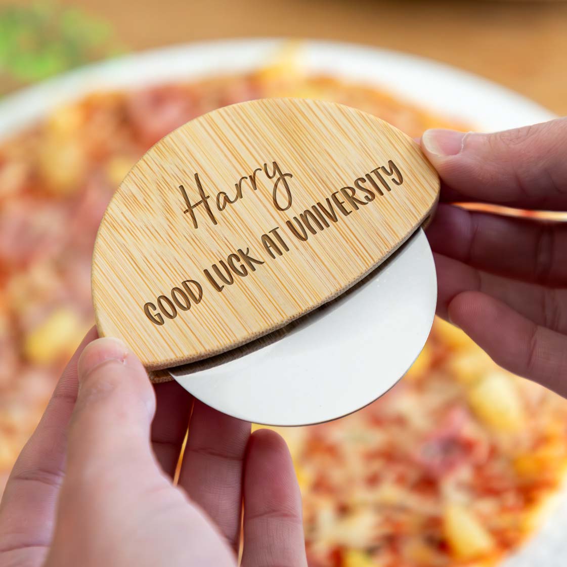 Wooden pizza cutter engraved with a personalised name and 'Good Luck at University' text held over a pizza.