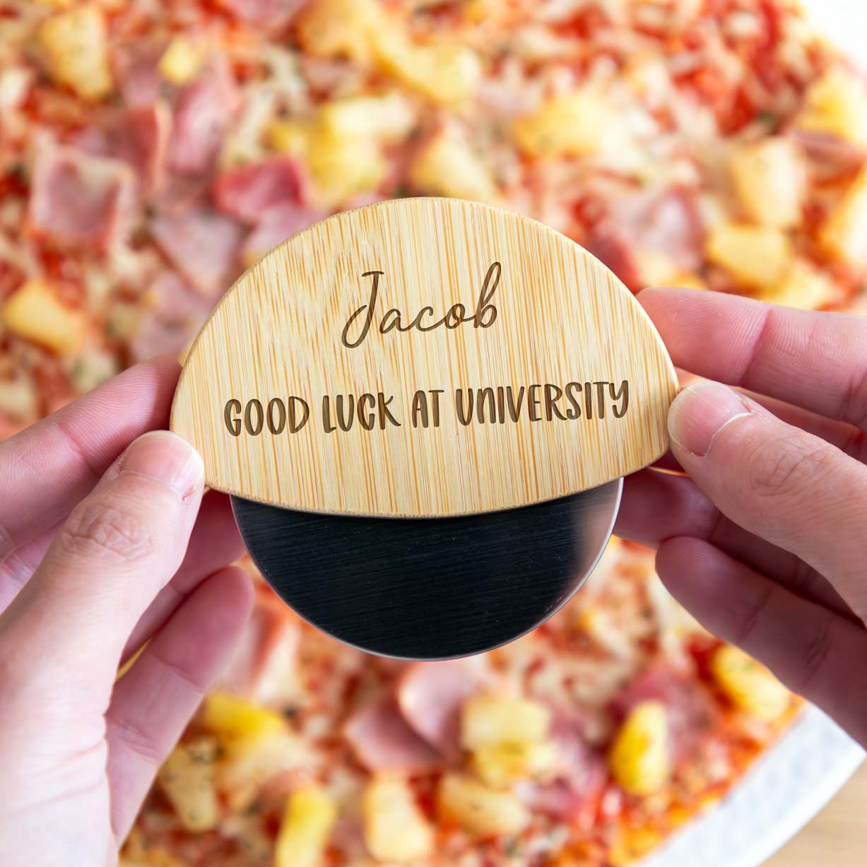 Person holding a wooden pizza cutter with engraved text in front of a pizza.
