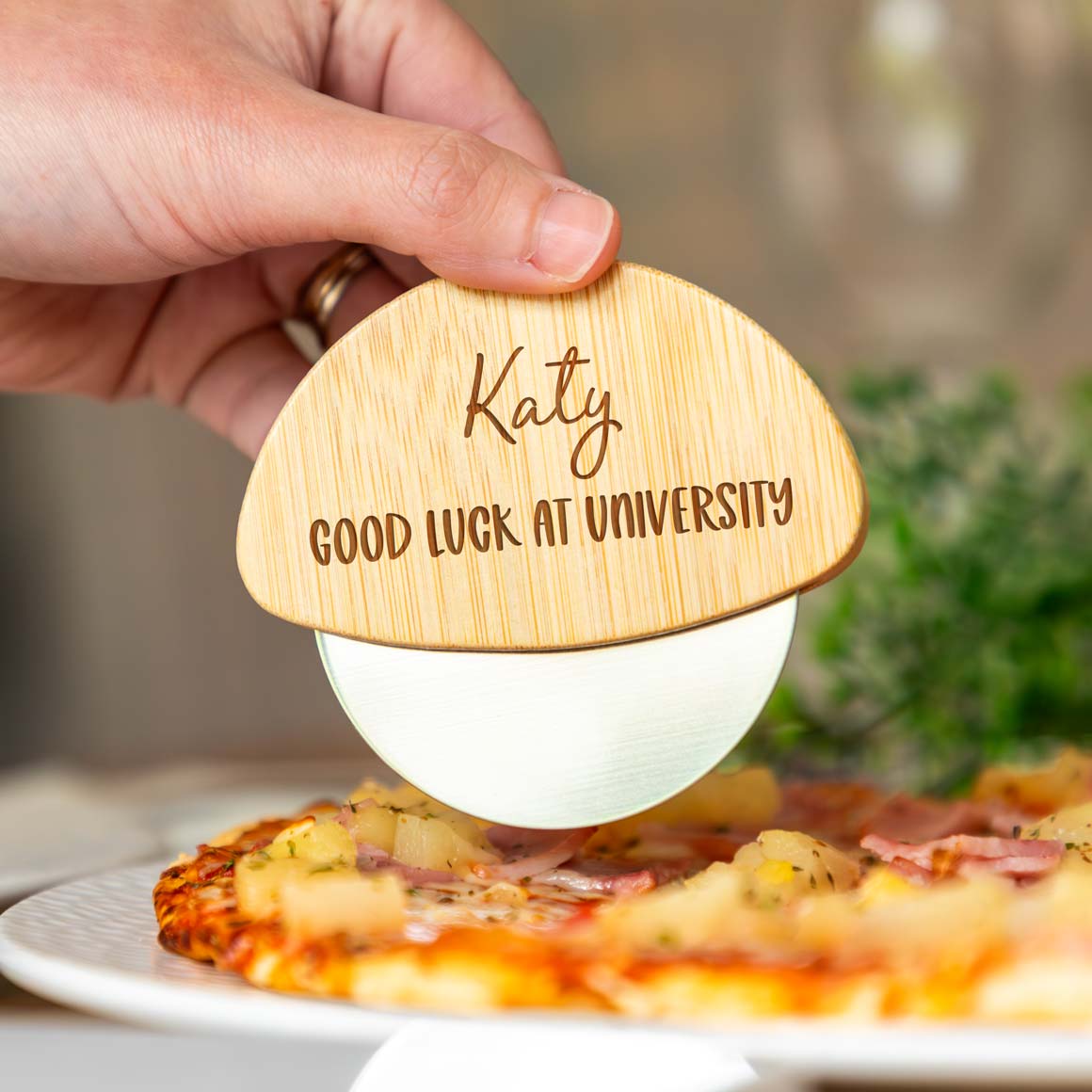 Wooden pizza cutter wheel engraved with a name and 'Good Luck at University' text over a pizza.
