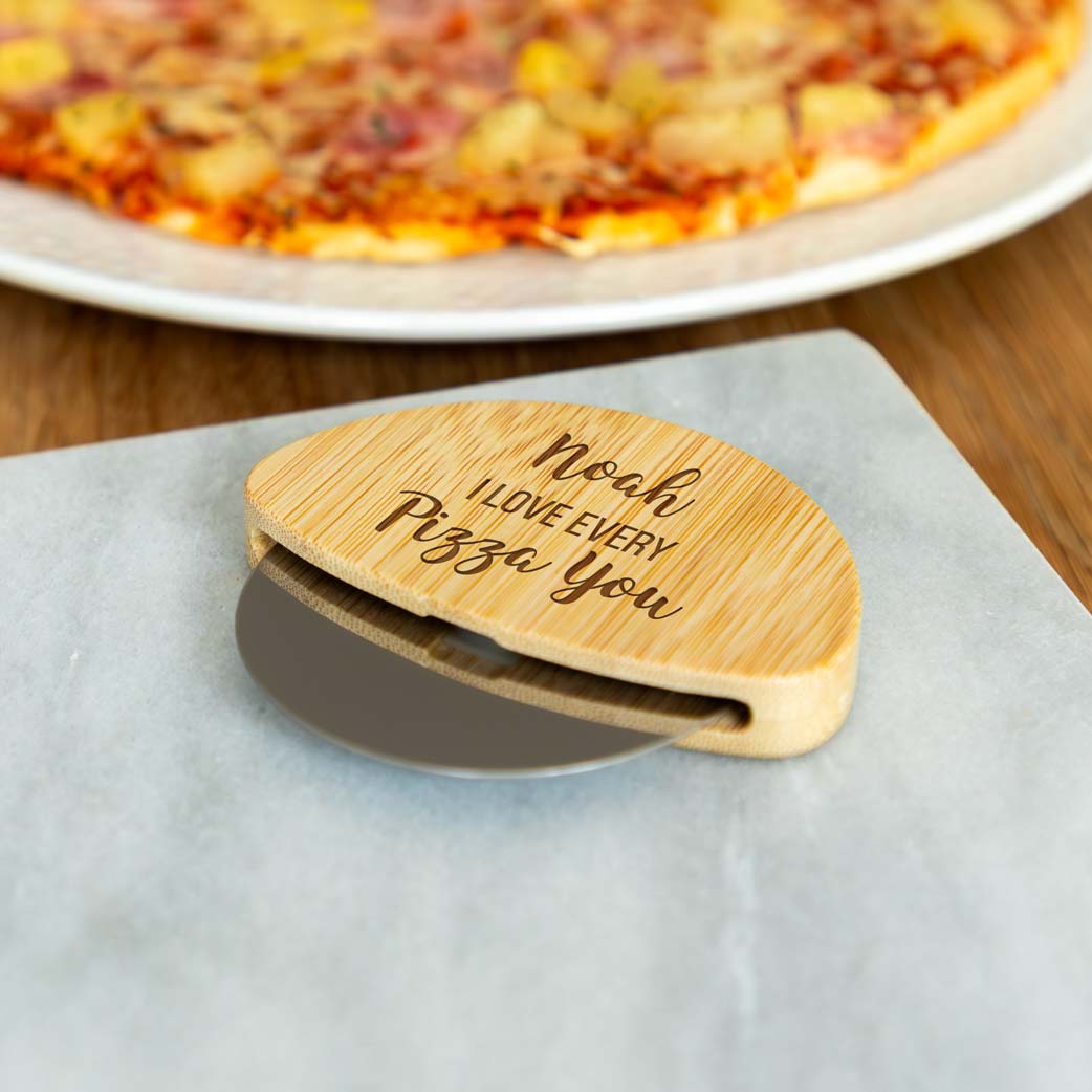 Wooden pizza cutter with a name and  'I love every pizza you' engraving on a table with a pizza in the background.