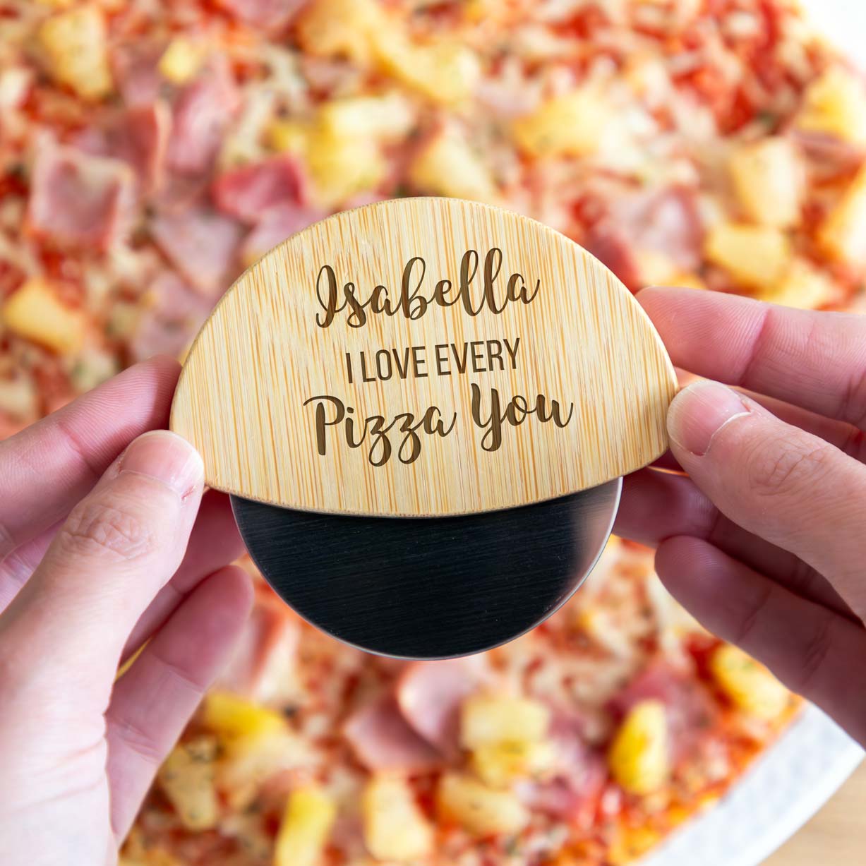 Wooden pizza cutter with engraved personalised text held over a pizza.