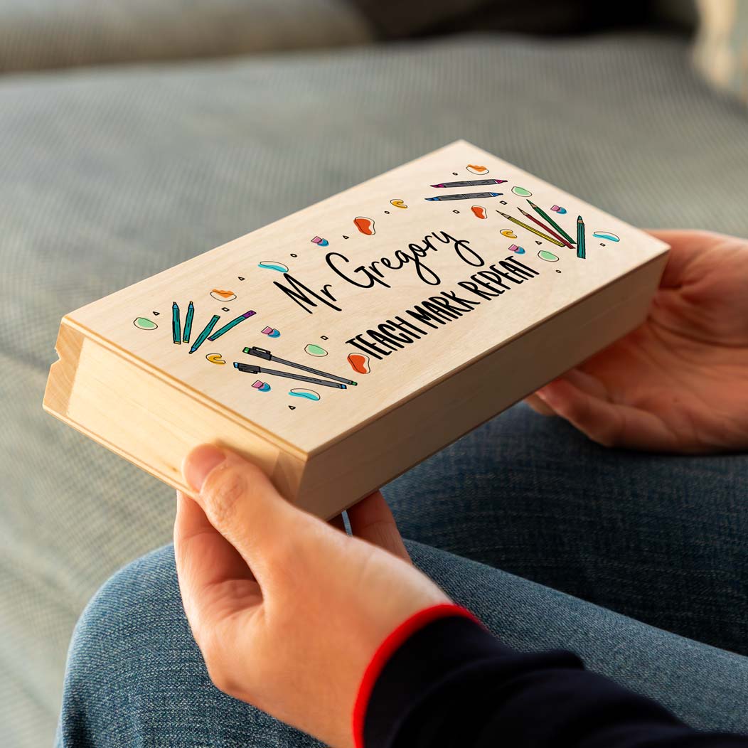 Person holding a wooden pencil case printed with colourful illustrations and 'teach mark repeat', personalised with a name.