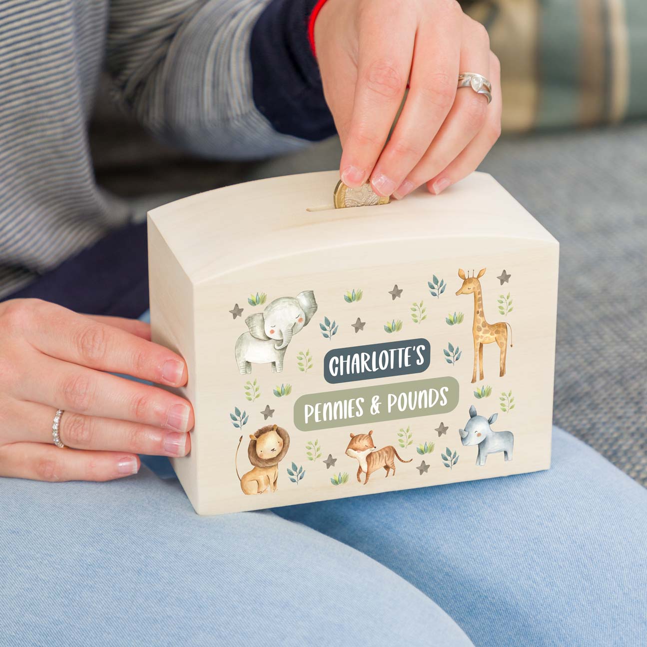 Wooden children's money box with jungle animal illustrations and personalised text, being used by a person to put a coin inside.