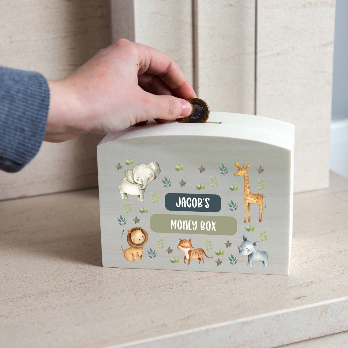 Hand placing a coin into a children's money box with jungle animal illustrations and personalised text.