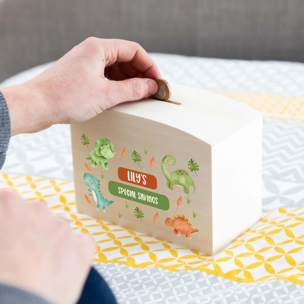 Person depositing money into a children's savings box with dinosaur design and personalised text.