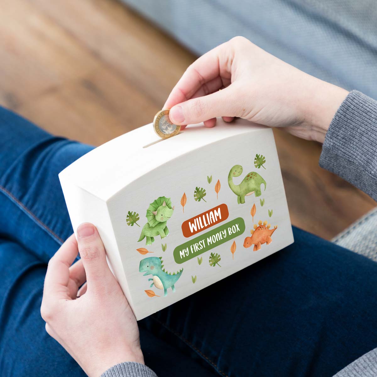 Person depositing a coin into a children's money box with dinosaur illustrations and a personalised name with 'My First Money Box'.