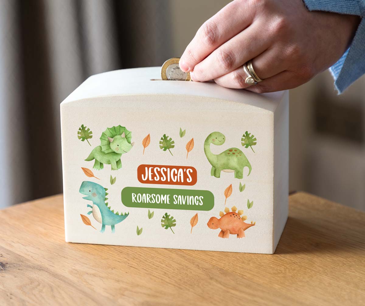 Personalised dinosaur-themed savings box with a hand placing a coin into it on a wooden surface.