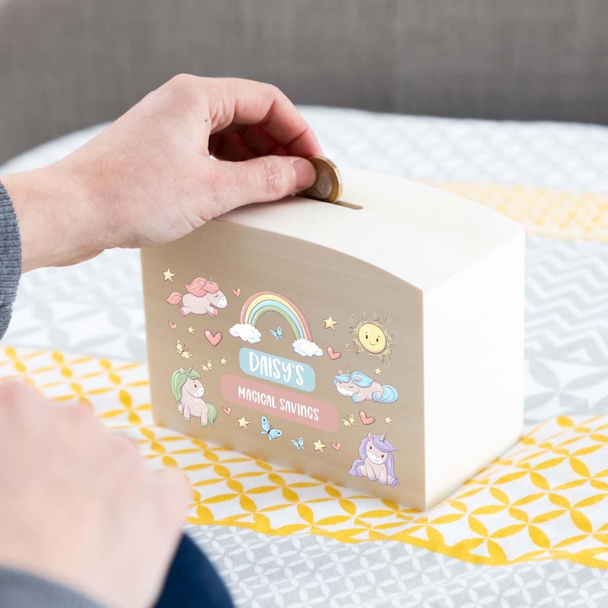 Person depositing money into a personalised children's savings box with unicorn and rainbow designs.