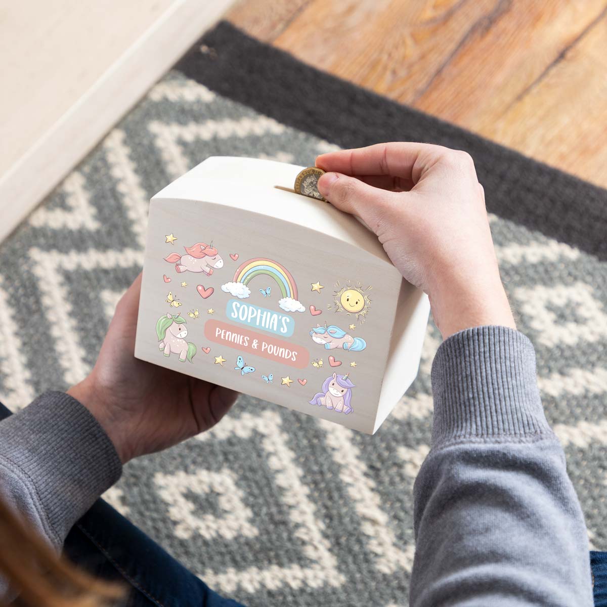 Person depositing a coin into a personalised money box with unicorn designs and personalised text.