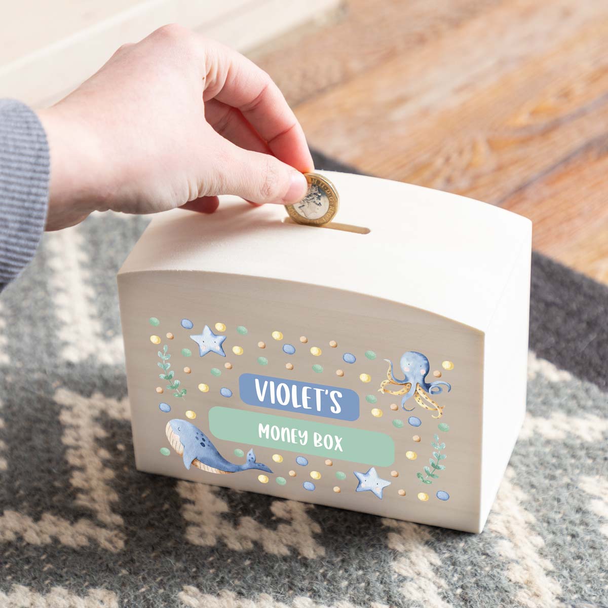Hand placing a coin into a personalised children's money box with a sea animal design.