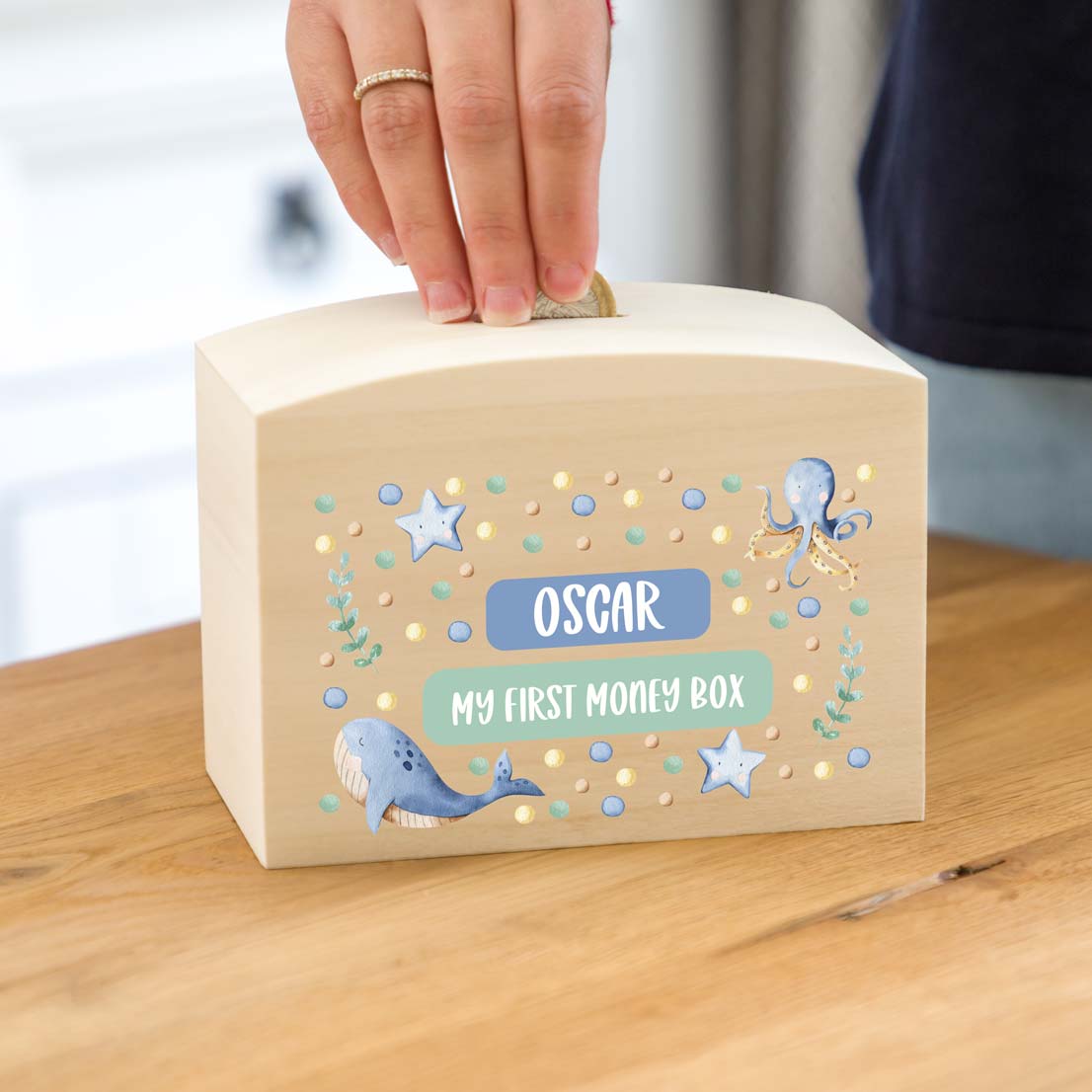 Wooden money box with a personalise name and 'My First Money Box' text, decorated with sea-themed illustrations on a wooden surface.
