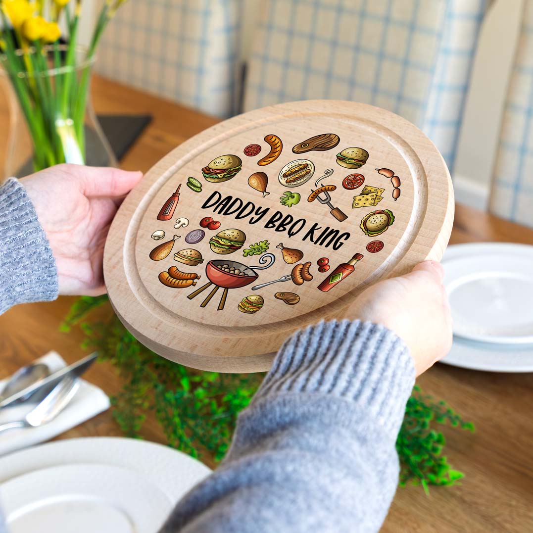 Person holding a wooden serving board with 'Daddy BBQ King' personalised text and grill illustrations on a dining table.
