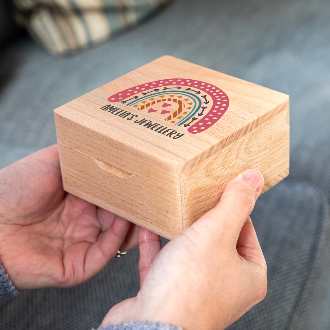 Wooden jewellery box held in two hands, with a colourful patterned rainbow and personalised text printed on the lid.