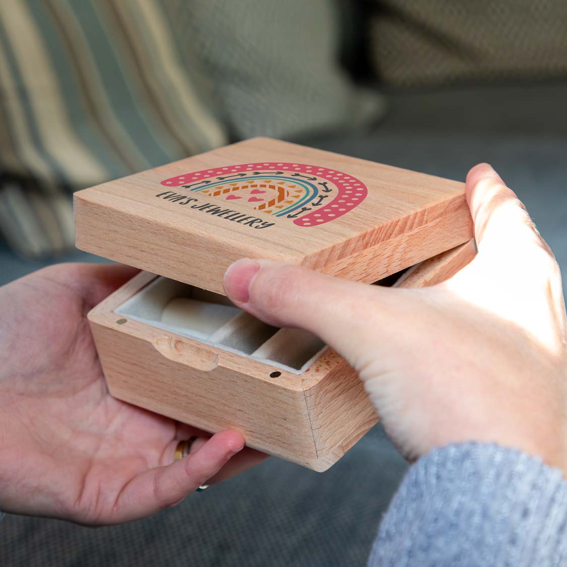 Hands lifting the personalised rainbow lid of a wooden jewellery box, revealing cream-lined storage compartments.