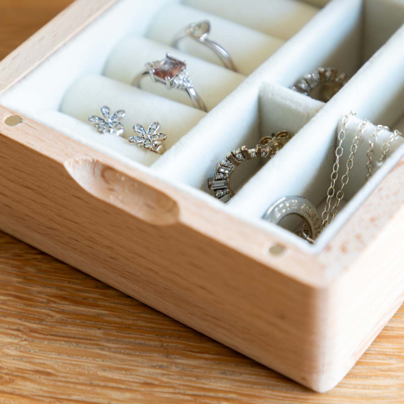 Close-up of cream-lined compartments in a wooden jewellery box, holding rings, flower earrings and necklaces.