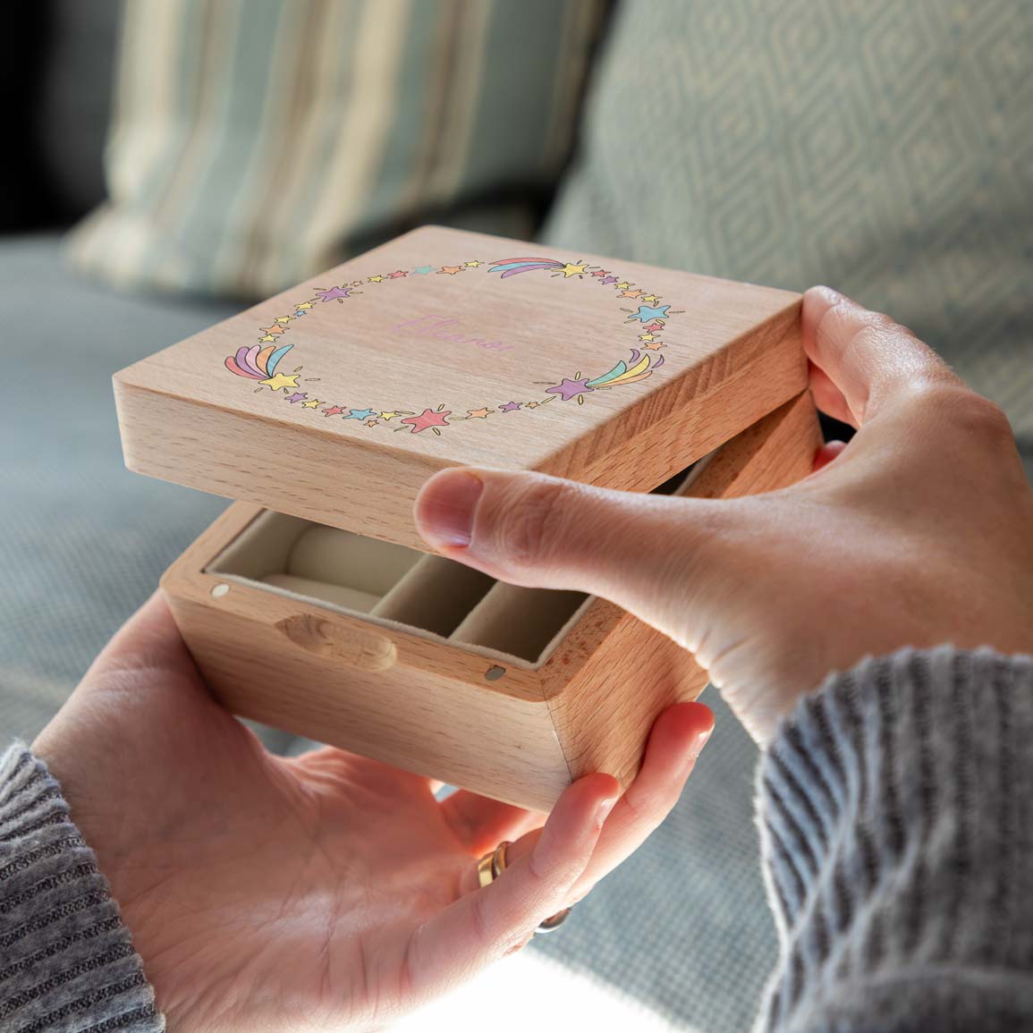 Hands lifting the shooting-star lid of a wooden jewellery box, revealing empty cream-lined compartments.
