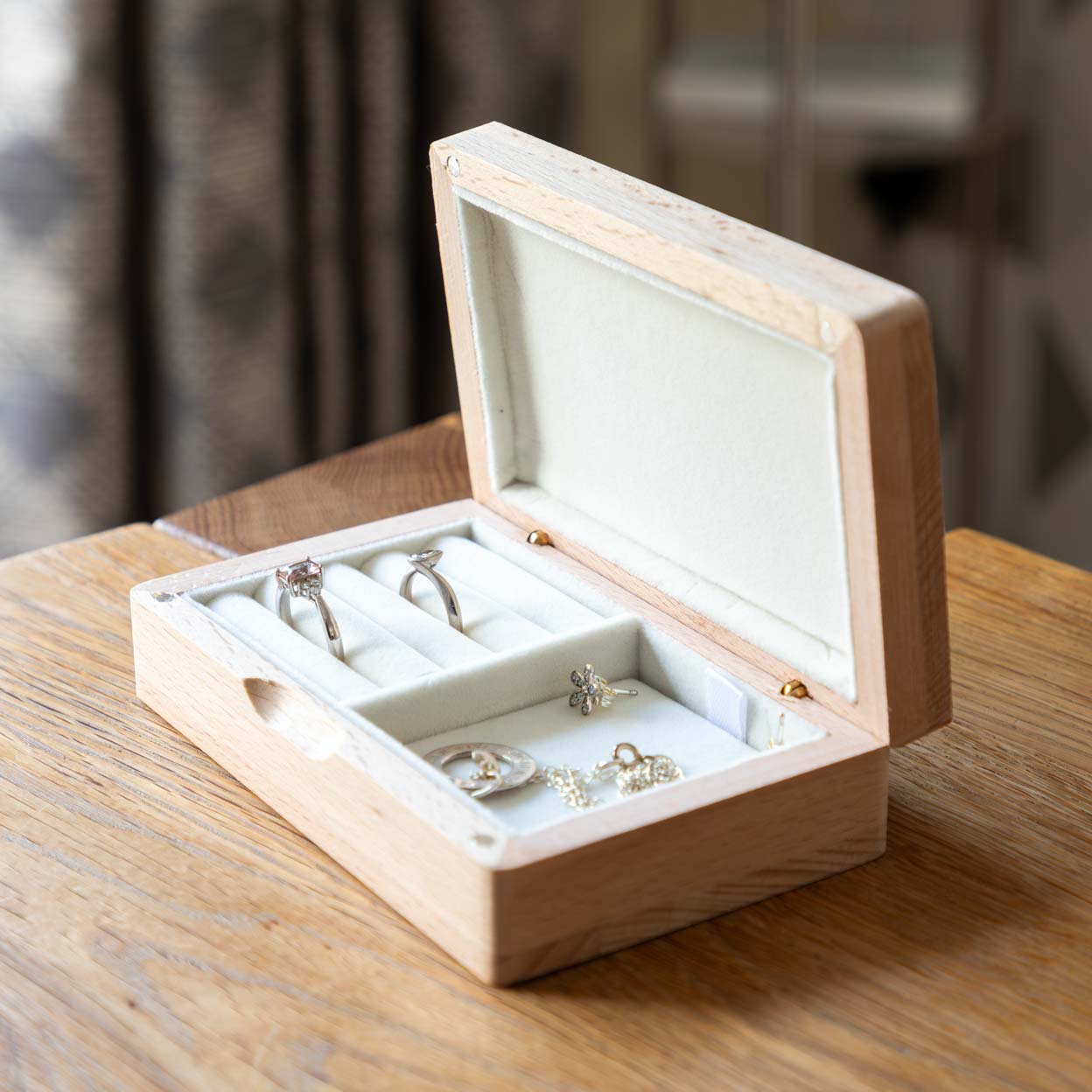 Open wooden jewellery box on a table, with two rings in cream-lined rolls and jewellery in the side compartment.