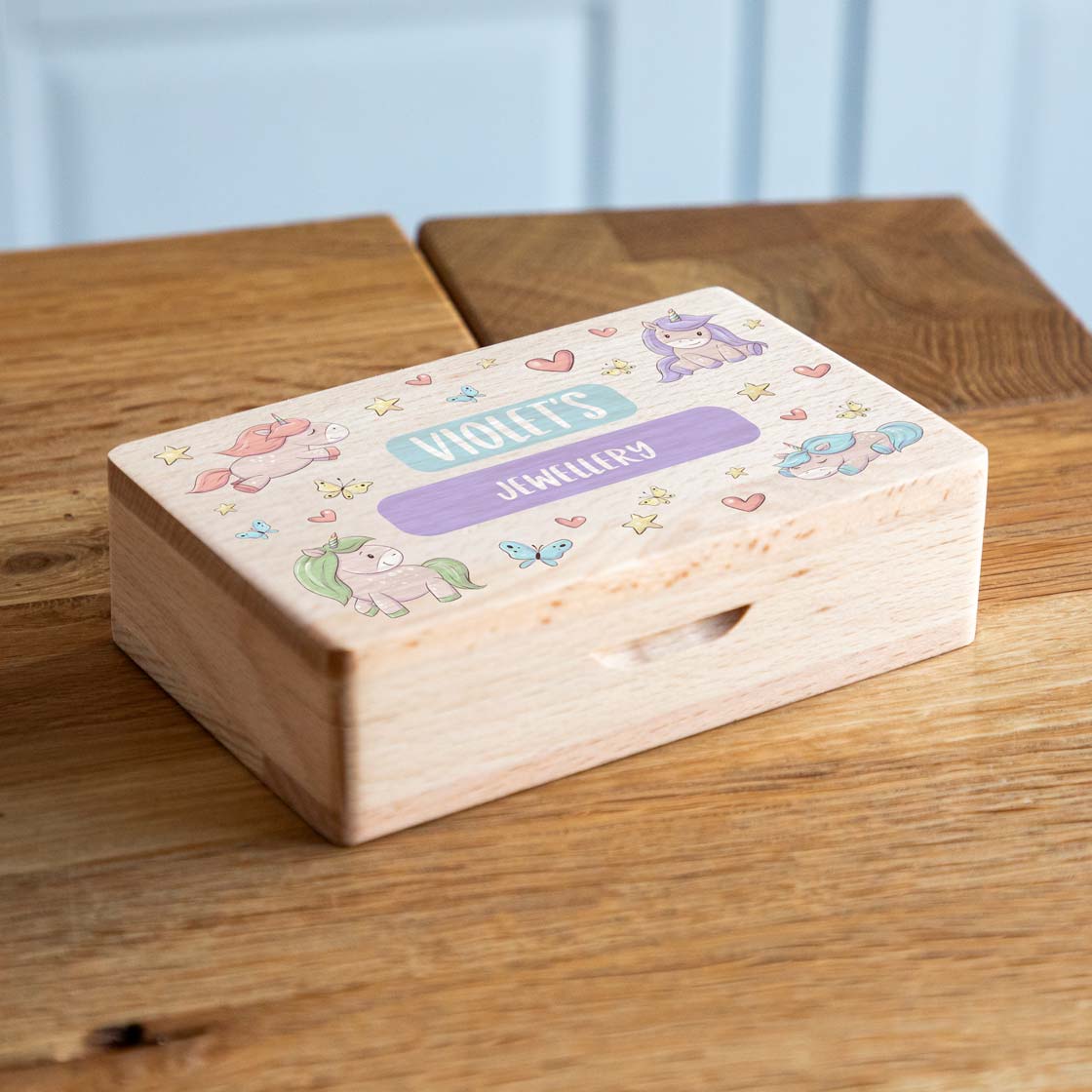 Wooden jewellery box on a table, with colourful unicorns, hearts and stars surrounding personalised text on the lid.
