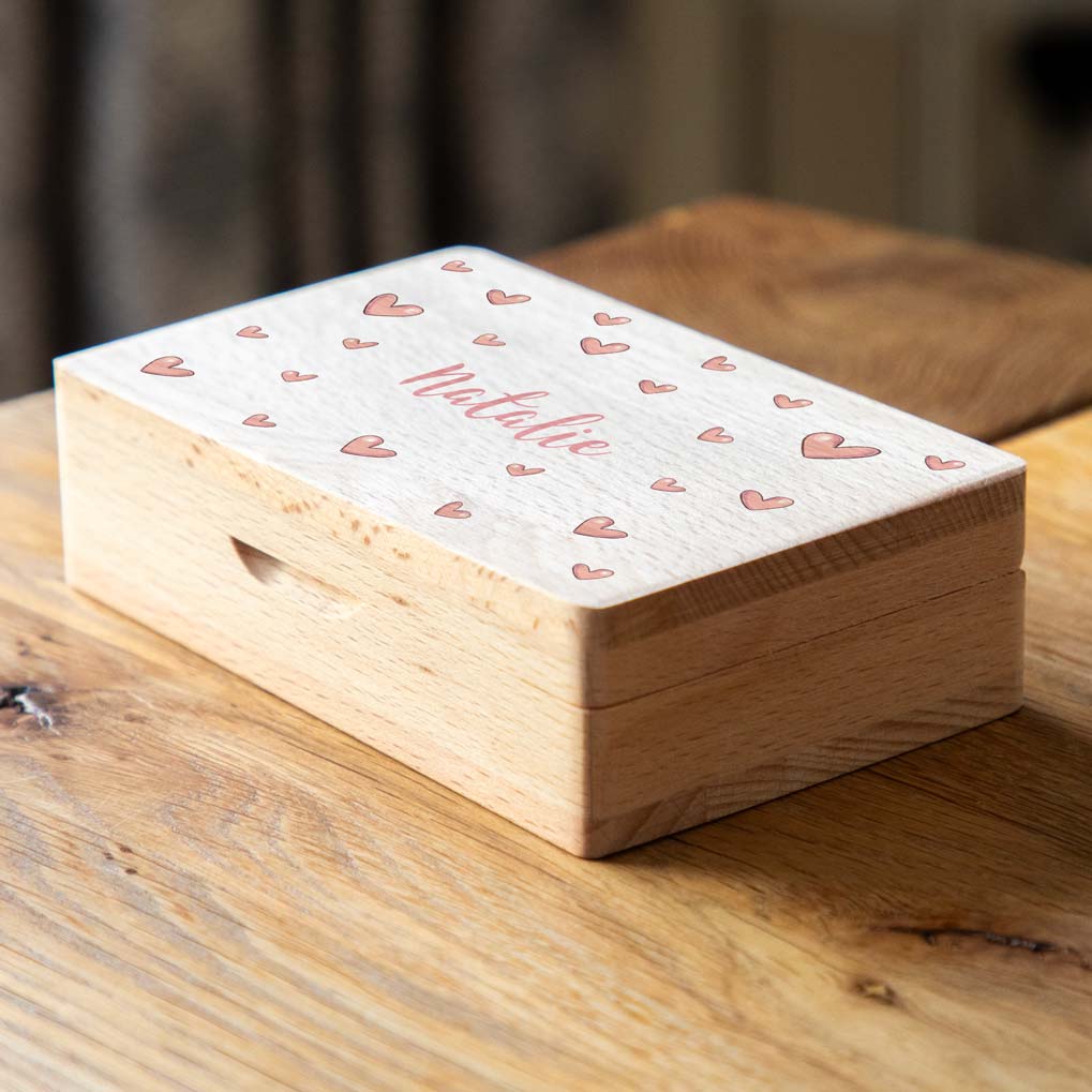 Closed wooden jewellery box on a table, with pink hearts surrounding the custom name on the lid.